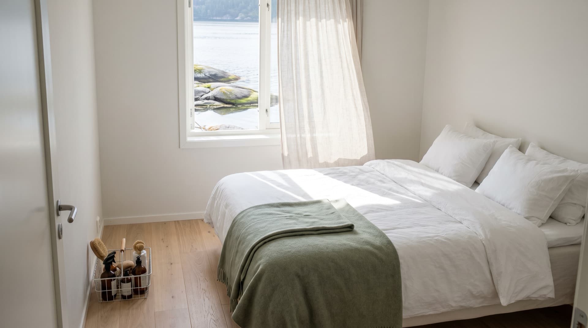 Freshly made bed with white linen and a sage throw in a bright rental bedroom overlooking the fjord, cleaning caddy by the door after turnover cleaning